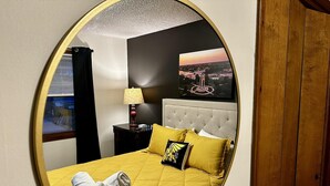 Ein Hotelzimmer mit einem großen, ovalen Spiegel, einem Bett mit gelber Bettwäsche, einem Kopfteil, einem Nachttisch mit einer Lampe, einem gerahmten Bild an der Wand und einem Holzschrank.