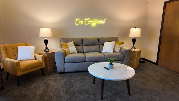 Ein gemütliches Wohnzimmer mit einem grauen Sofa, zwei Sesseln, einem runden Couchtisch mit einer Pflanze und zwei Lampen. Ein Neon-Schild darüber zeigt den Schriftzug "Go Griffins!".
