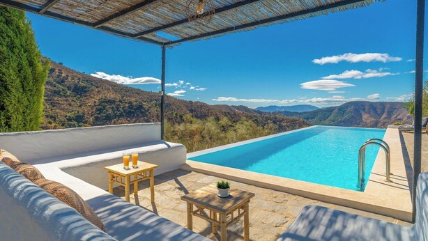 Zona junto a la piscina con vistas a montañas, una mesa de madera con bebidas y un área de descanso acolchada.