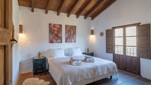 Un dormitorio con cama, dos cuadros en la pared, una mesita de noche con lámpara, una puerta de madera y un ventanal con contraventanas.