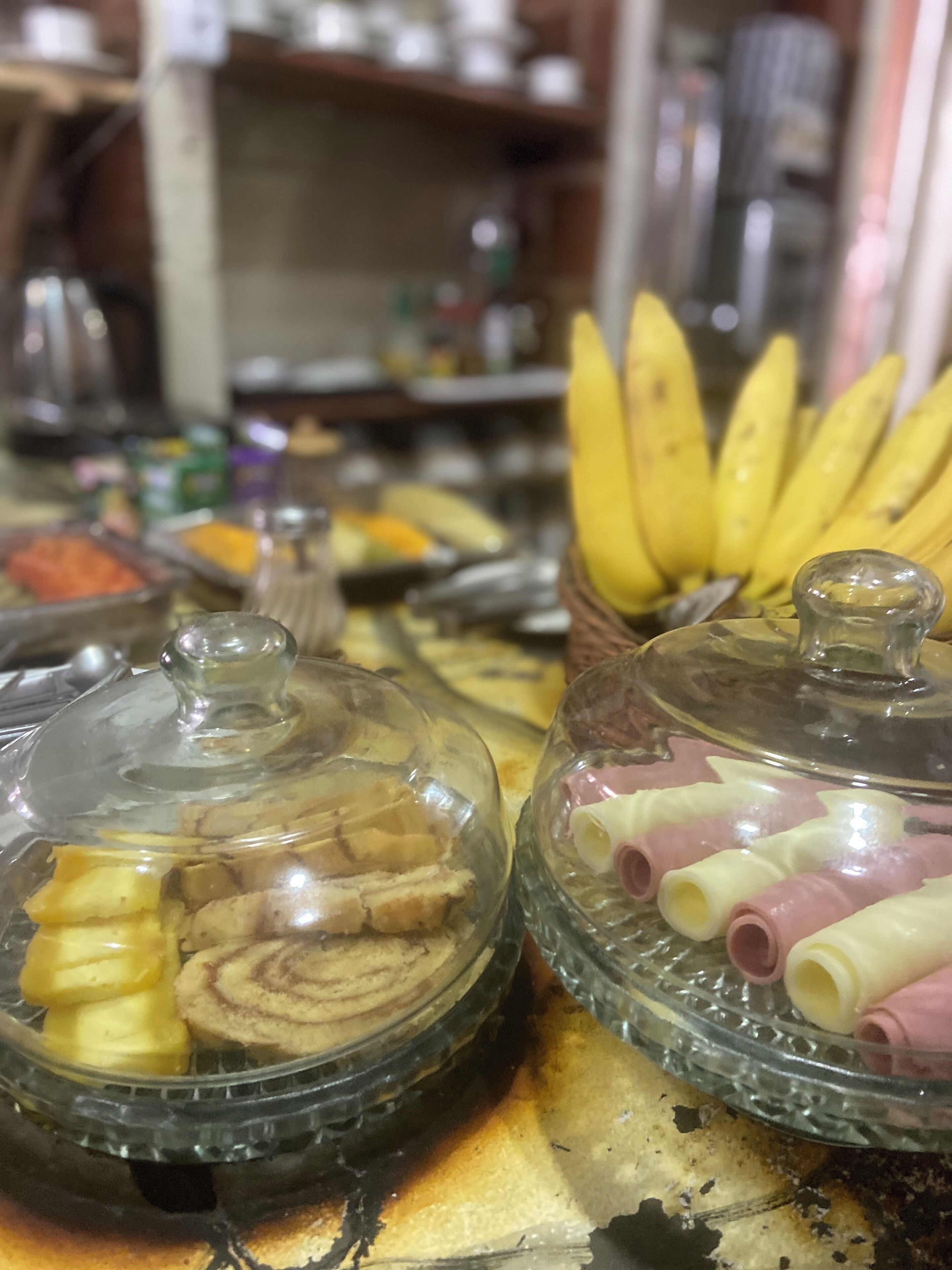 Un bufé con diversos alimentos bajo campanas, incluyendo pasteles y plátanos.