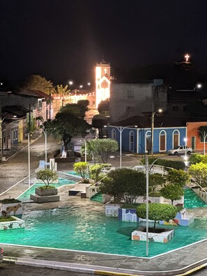 Vista nocturna de una plaza con una fuente, faroles y el campanario de una iglesia al fondo.