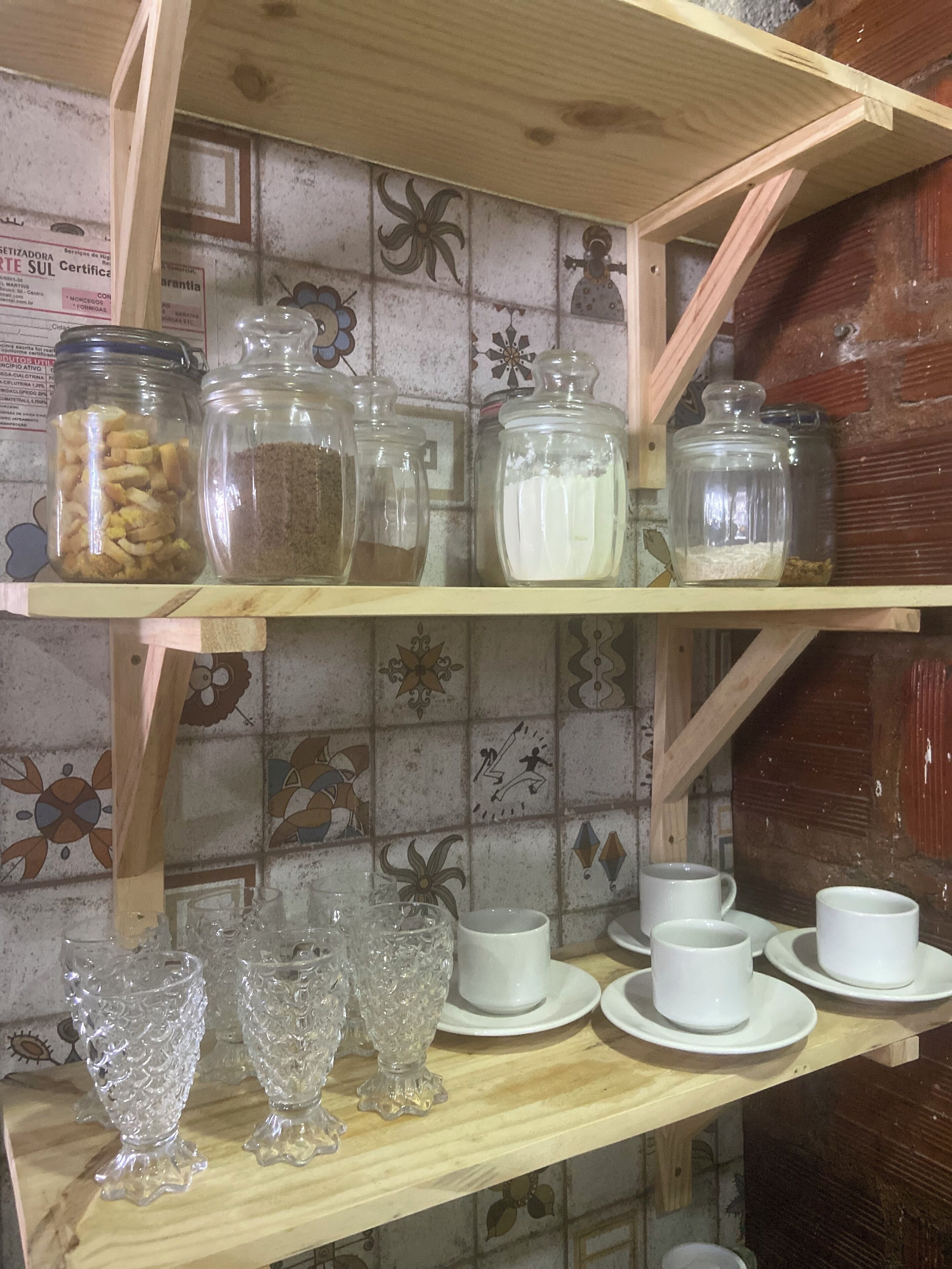 Una repisa en la cocina con frascos de vidrio que contienen diversos alimentos, un juego de tazas y platillos blancos, y cristalería decorativa.