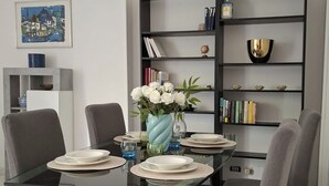 Una zona de menjador moderna amb una taula de vidre, cadires grises i una biblioteca amb llibres i objectes decoratius.