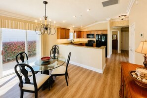 A modern kitchen with a central island, wooden cabinets, and a dining area with a glass table and chairs. - Sunshine Stay a Creekside Villa at Lake Sumter Landing with Nature & Water Views (The Villages)