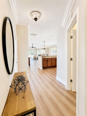 A modern kitchen with wooden cabinets and a central island, a dining area with a wooden table, and a hallway with a large mirror and a chandelier.