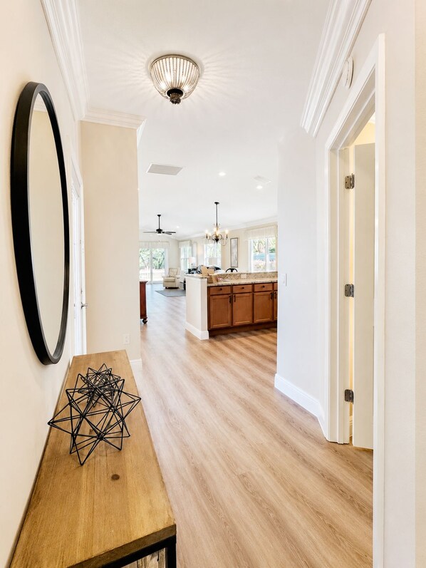 A modern kitchen with wooden cabinets and a central island, a dining area with a wooden table, and a hallway with a large mirror and a chandelier.