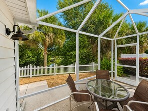 A covered patio with a glass table and chairs, surrounded by a white fence and lush greenery.