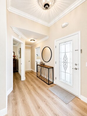 A hallway with wooden flooring, a console table with rope accents, a round mirror, and a door with a geometric glass design.