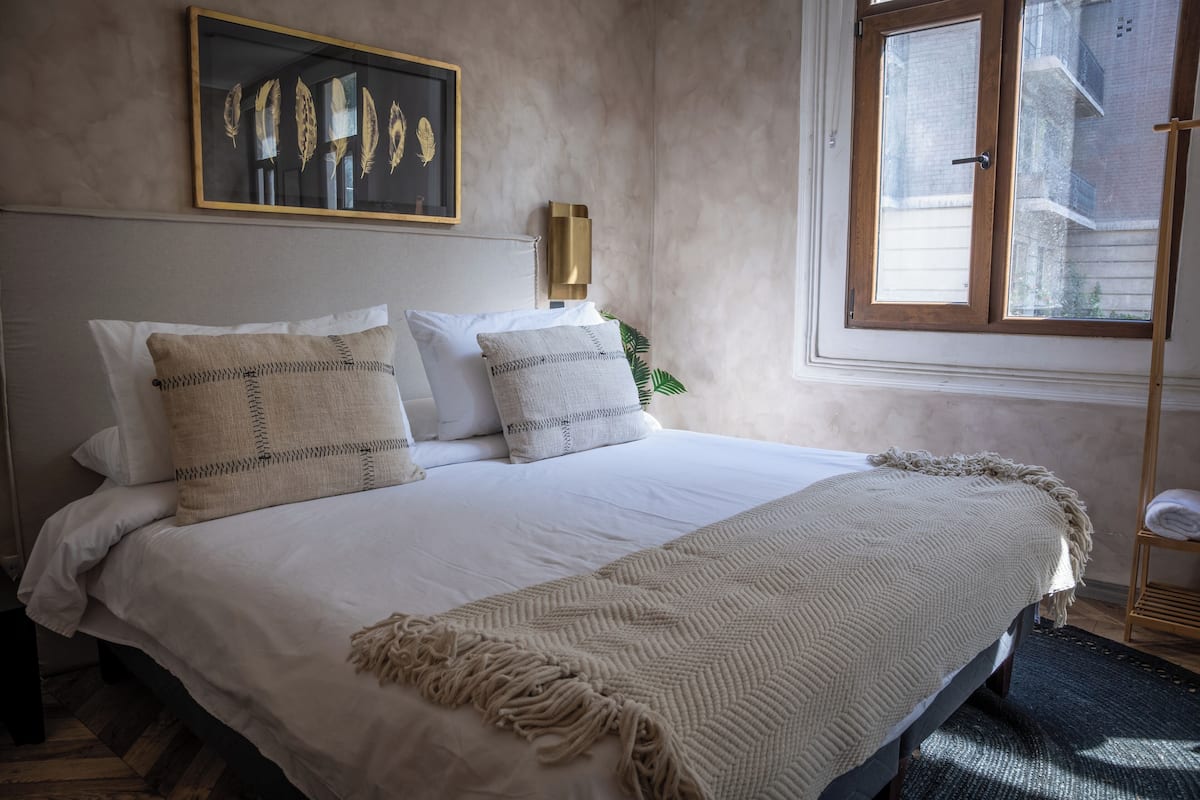 A neatly made bed with a white comforter and two decorative pillows, a framed artwork above the bed, and a window with a view of buildings.