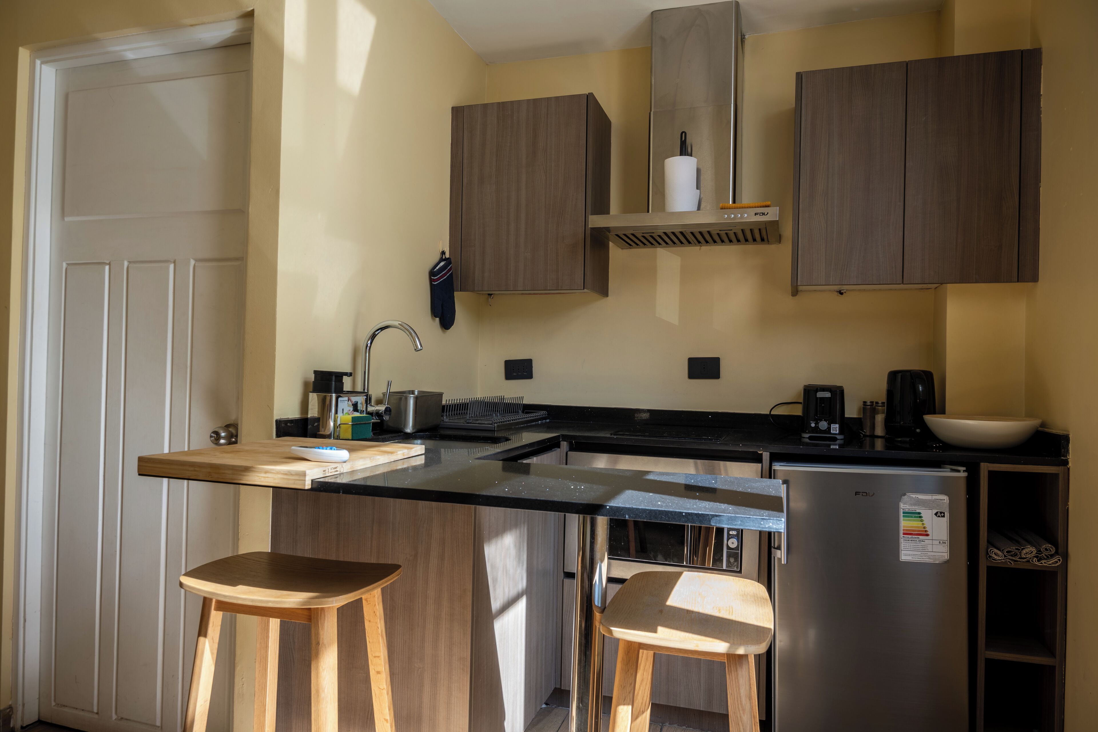 A compact kitchen with wooden cabinets, a stainless steel refrigerator, a coffee maker, and two wooden stools.
