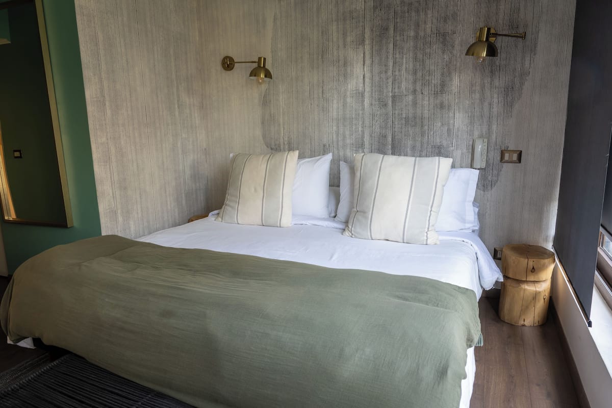 A neatly made bed with white linens and striped pillows, a wooden nightstand, and a textured wall with two wall-mounted lamps.