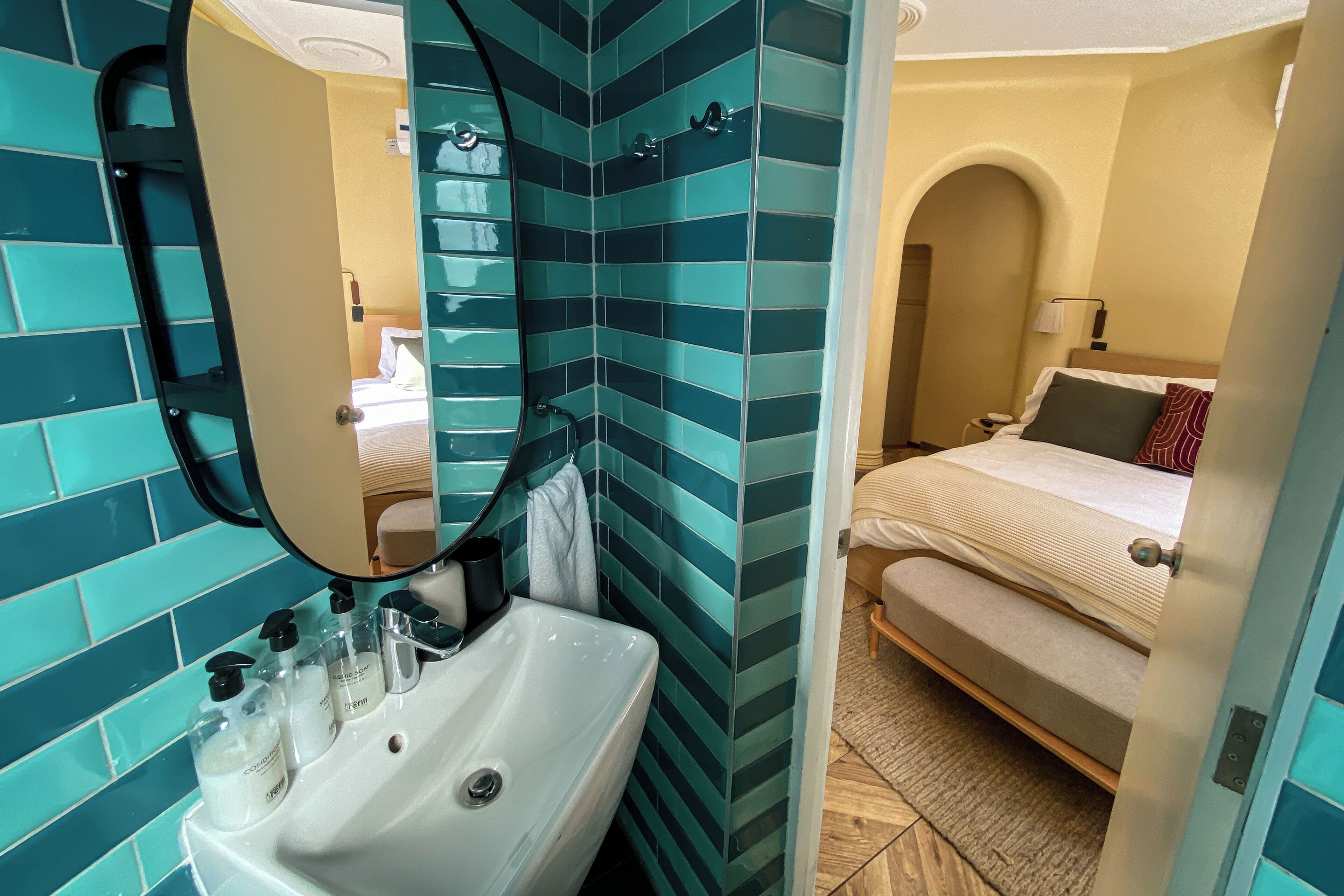 Une salle de bain équipée d’un lavabo, d’un miroir et d’articles de toilette. On peut apercevoir, dans la pièce adjacente, un lit avec une table de chevet et une lampe.
