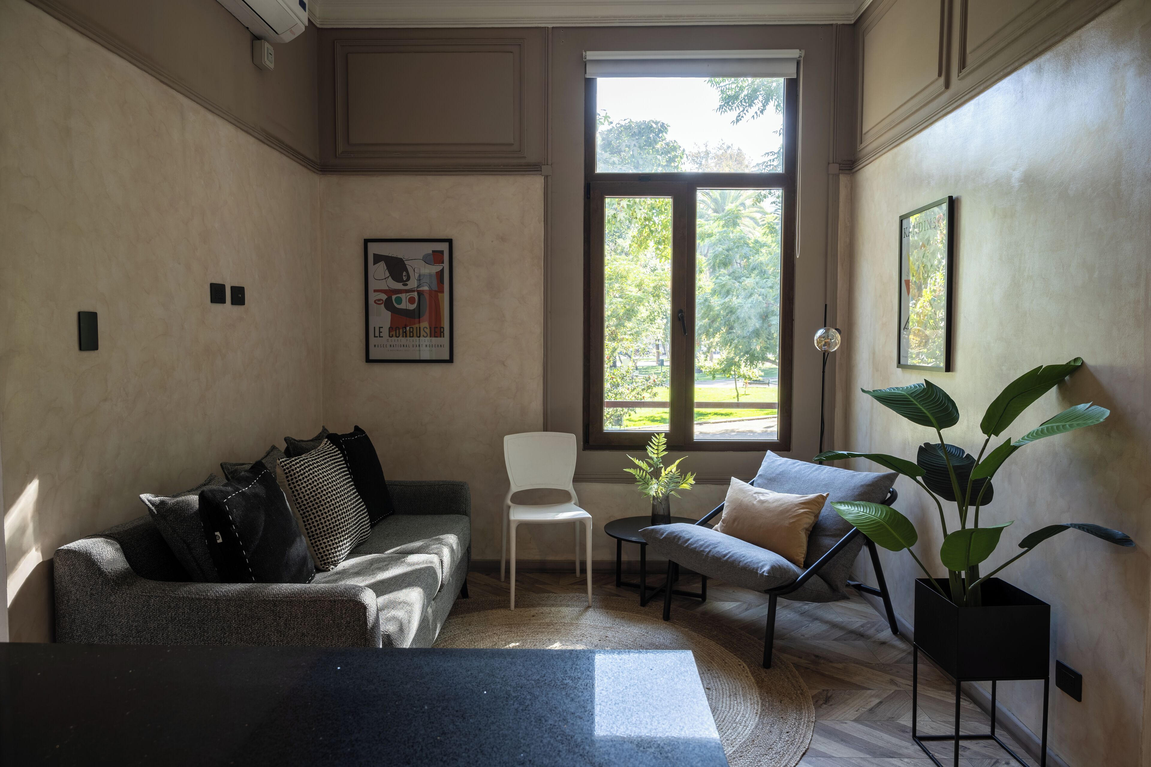 A modern living room with a grey sofa, a white chair, and a black coffee table. There is a large window providing natural light and a view of greenery outside.