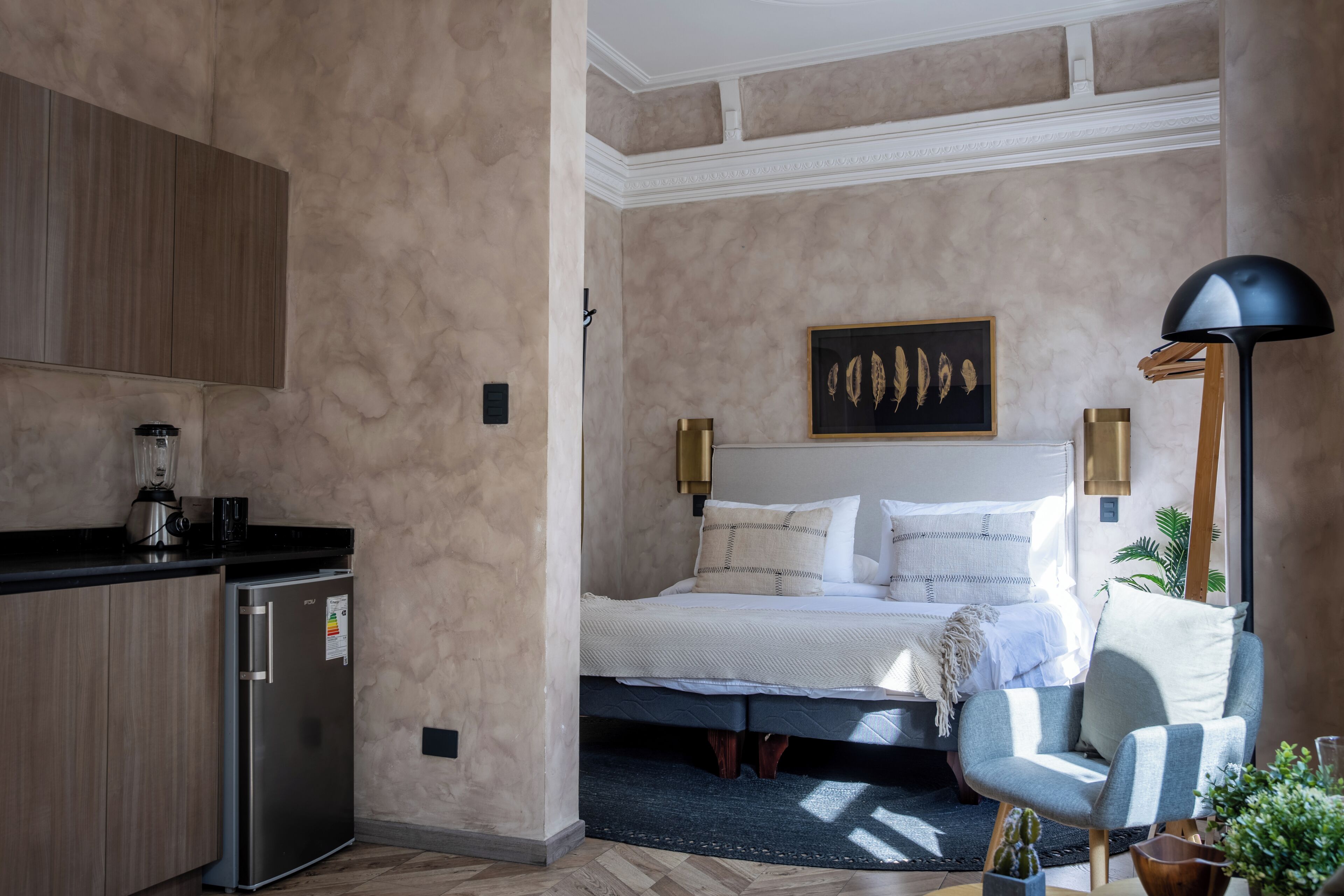 Un dormitorio con cama, mesita de noche, lámpara y una pequeña kitchenette con refrigerador y cafetera.
