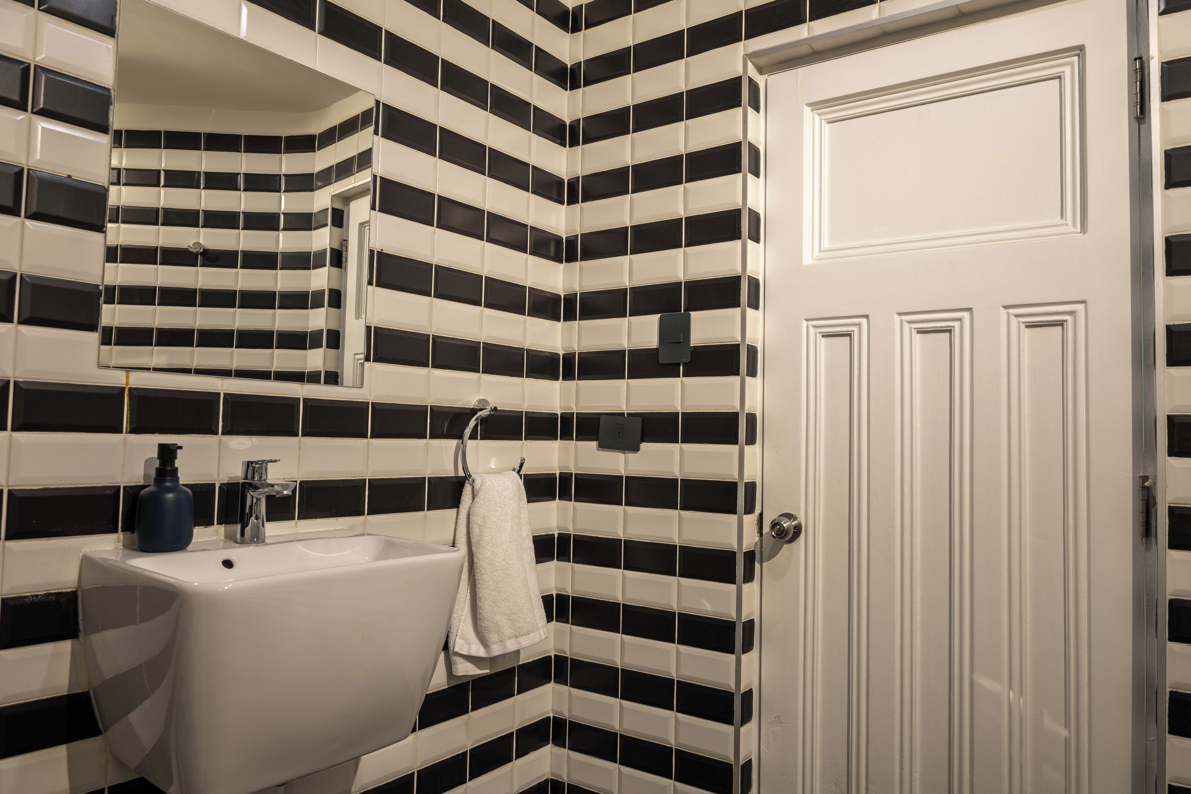 Ein Badezimmer mit weißem Waschbecken, schwarz-weißen Streifenfliesen, einem Spiegel und einer weißen Tür.