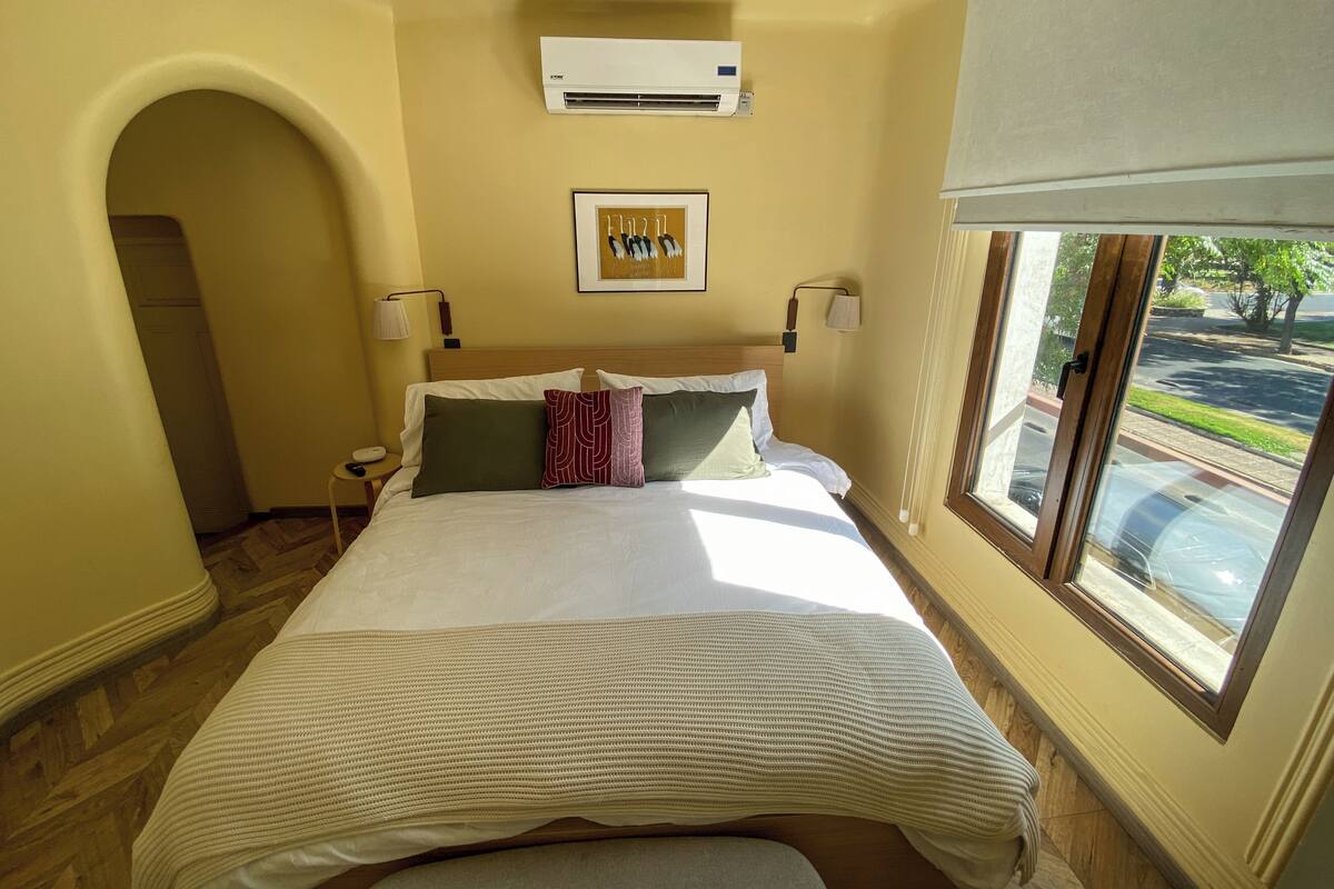 A hotel room with a bed, an air conditioner, a framed picture, a window, and a lamp.