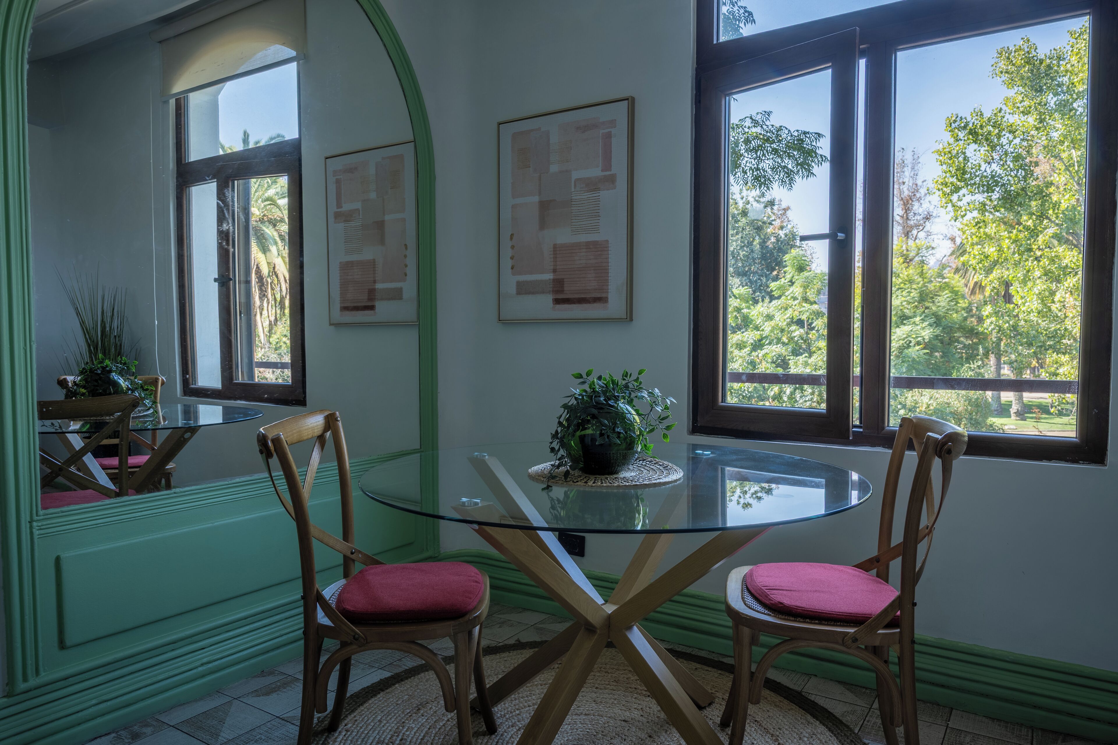 Een eethoek met een glazen tafel, twee houten stoelen met rode kussens, en een potplant op de tafel.