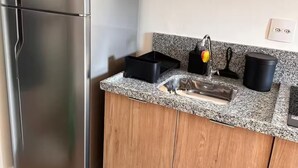 Una cocina compacta con un refrigerador de acero inoxidable, un microondas sobre un estante de madera y una encimera de granito con un fregadero y electrodomésticos de cocina negros.