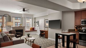 A modern living room with a sofa, coffee table, and a dining area with a table and chairs. There is a fireplace, a TV mounted on the wall, and a kitchen area with a microwave and stove.
