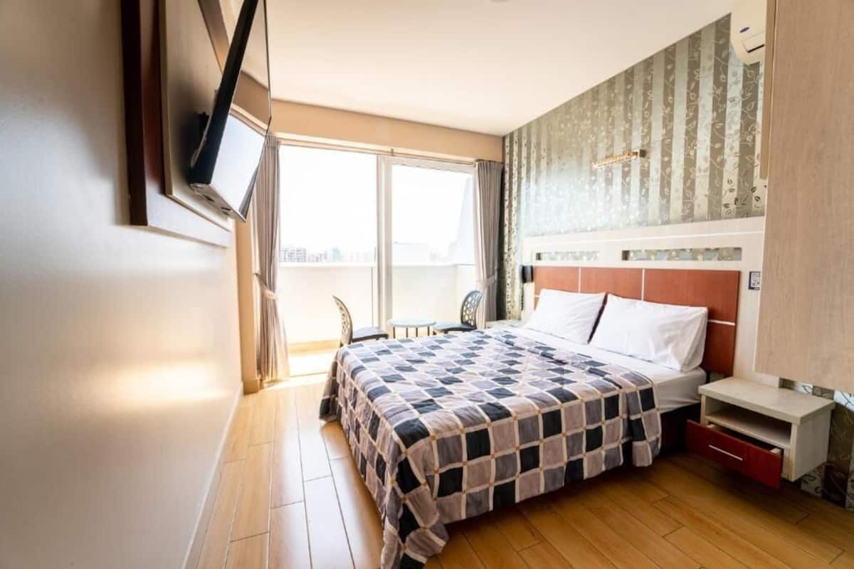 A hotel room with a large bed, a TV mounted on the wall, a small table, and a window with a view of the city.