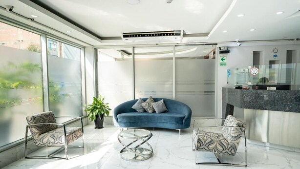 A modern hotel lobby with a blue sofa, two patterned armchairs, a glass coffee table, and a large window offering a view of greenery. - HOTEL QUADRUM (Lima)