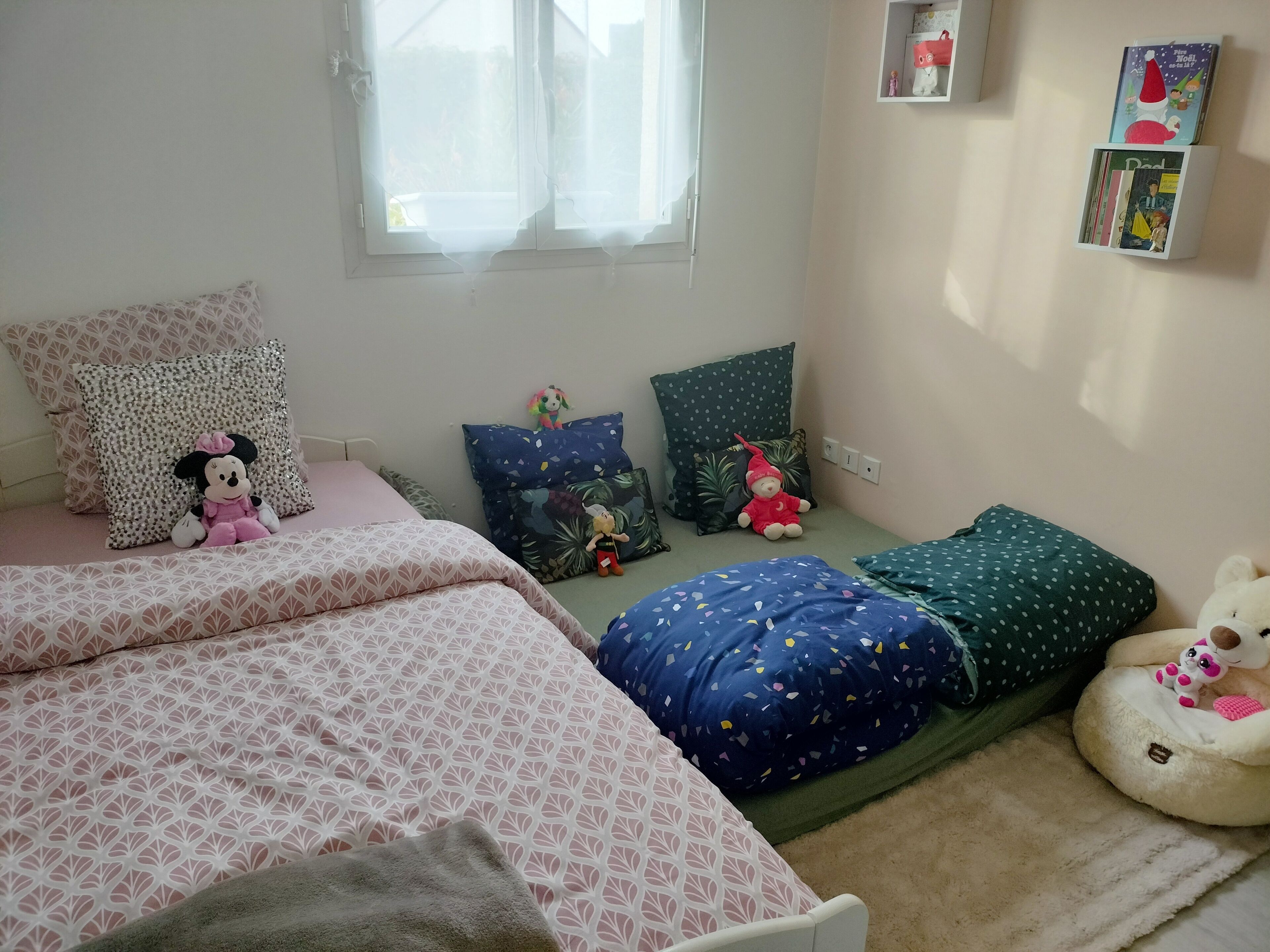 Een slaapkamer met een bed, kussens, een teddybeer en een plank met boeken.