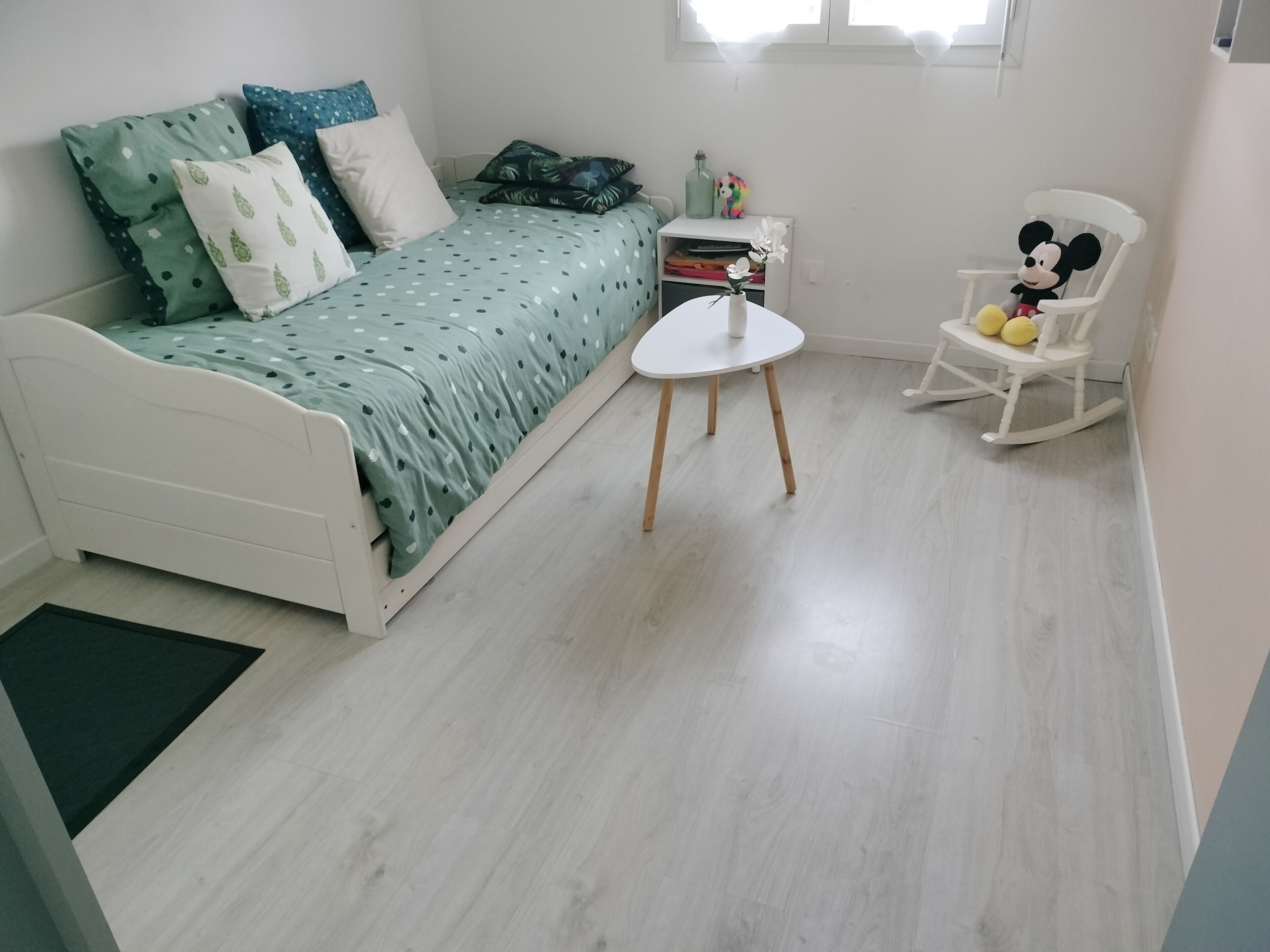 A bedroom with a bed, a small table, a rocking chair, and a window.