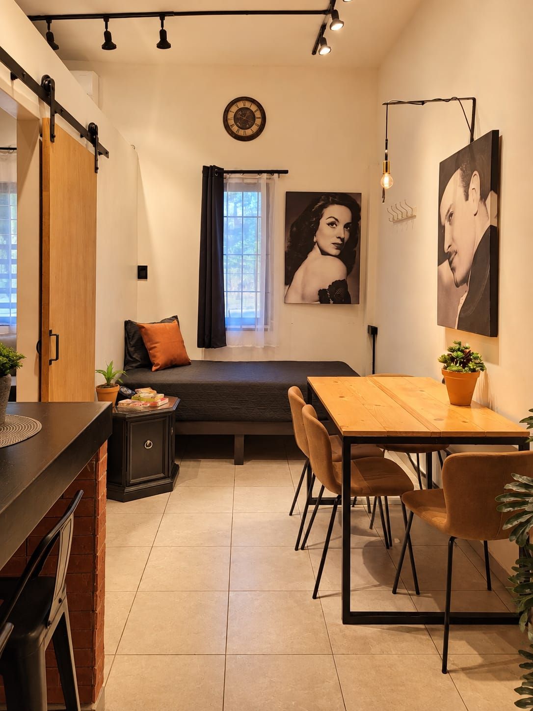 A compact room with a bed, a dining table with chairs, a small cabinet, and a window with curtains.