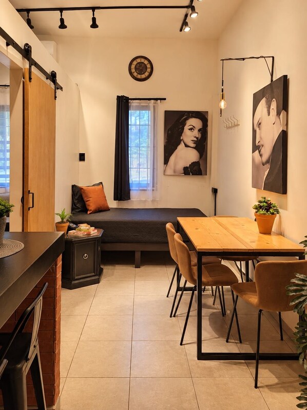 A compact room with a bed, a dining table with chairs, a small cabinet, and a window with curtains.