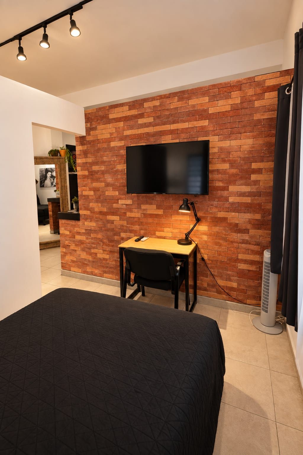 A modern room with a brick wall, a flat-screen TV mounted above a desk, a bed with a dark quilt, and a standing heater.