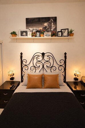 A bed with a black metal headboard, two bedside tables with lamps, a shelf above the bed with framed pictures and plants, and a wall-mounted clock.