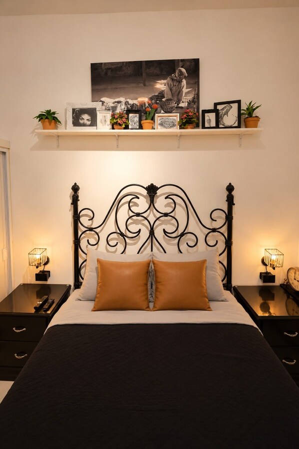 Un lit avec une tête de lit en métal noir, deux tables de chevet avec des lampes, une étagère au-dessus du lit ornée de cadres et de plantes, et une horloge fixée au mur.