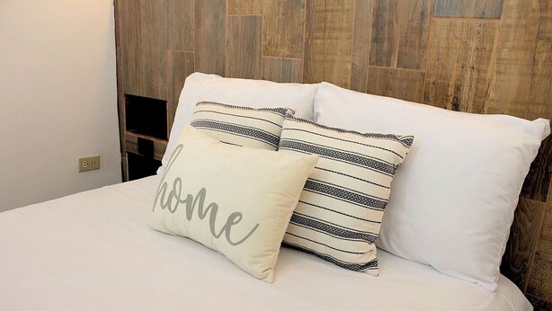 Een bed met witte beddengoed en twee kussens, waarvan er een het woord 'home' heeft.