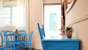 Une pièce avec une table bleue et des chaises assorties, un petit meuble bleu et une fenêtre donnant sur une porte bleue et un bâtiment.
