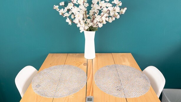 Una mesa de madera con un jarrón blanco lleno de flores blancas, dos sillas blancas y un control remoto sobre la mesa.