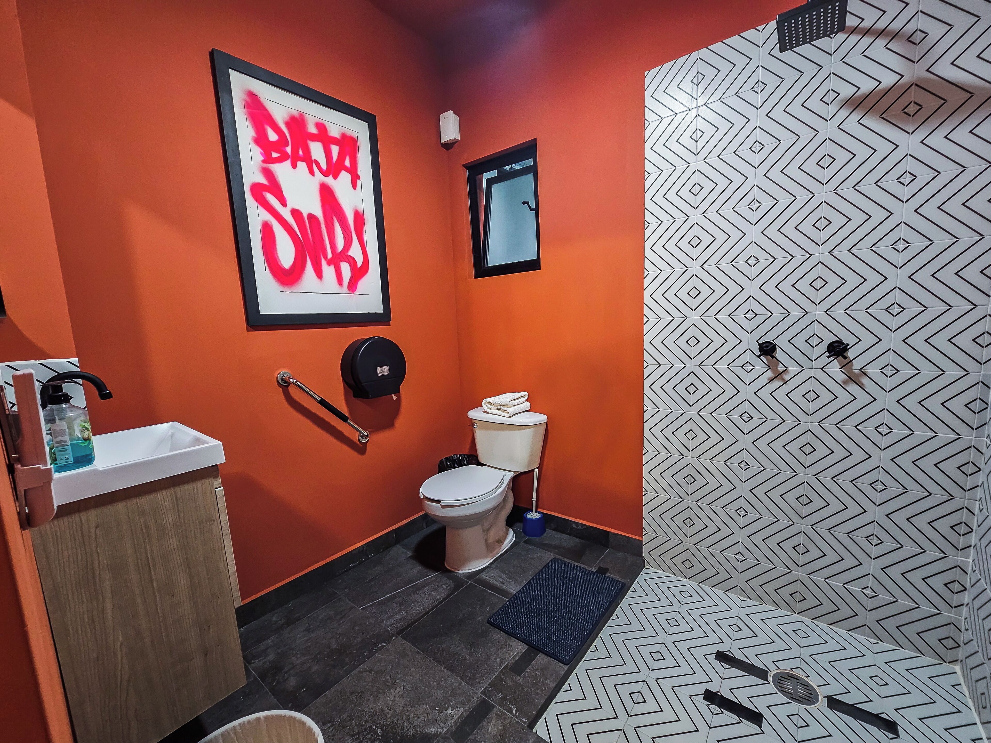 A bathroom with an orange wall, a toilet, a sink, a hand dryer, and a patterned shower wall.