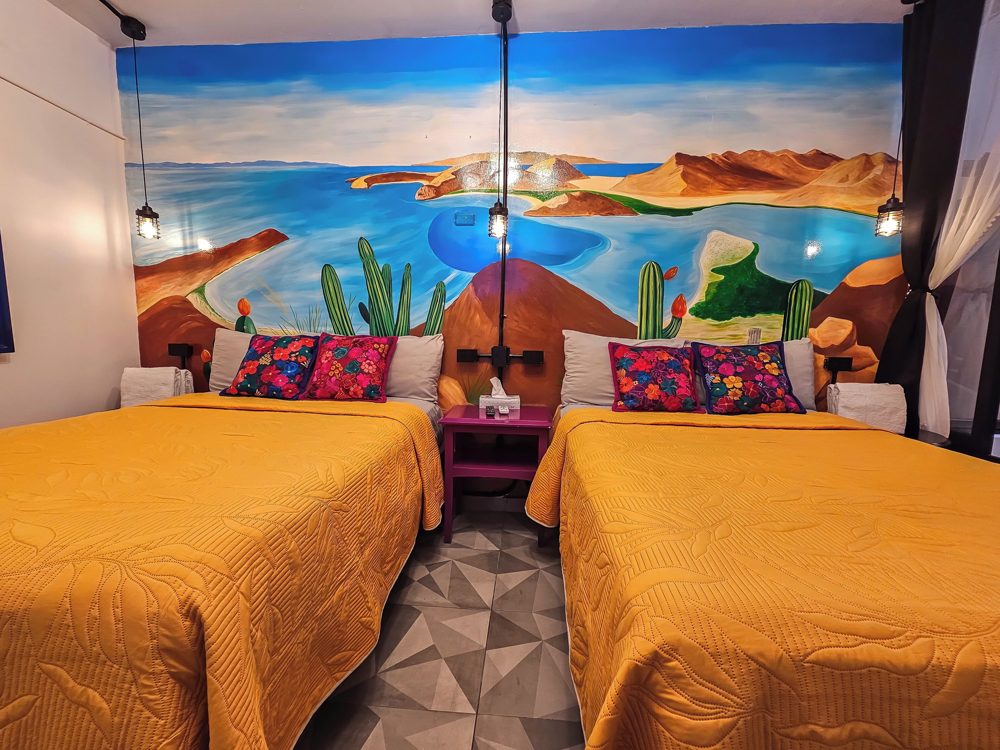 Una habitación con dos camas, cada una con colcha amarilla, un mural con paisaje desértico en la pared y un suelo con estampado geométrico.