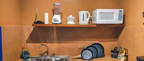 A kitchen with a sink, a microwave, a blender, and a kettle on a shelf.