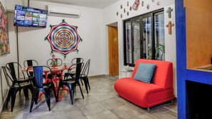 Ein Essbereich mit rotem Sofa, einem Fernseher, der einen Comedy-Sender zeigt, und einer farbenfrohen Wanddekoration.