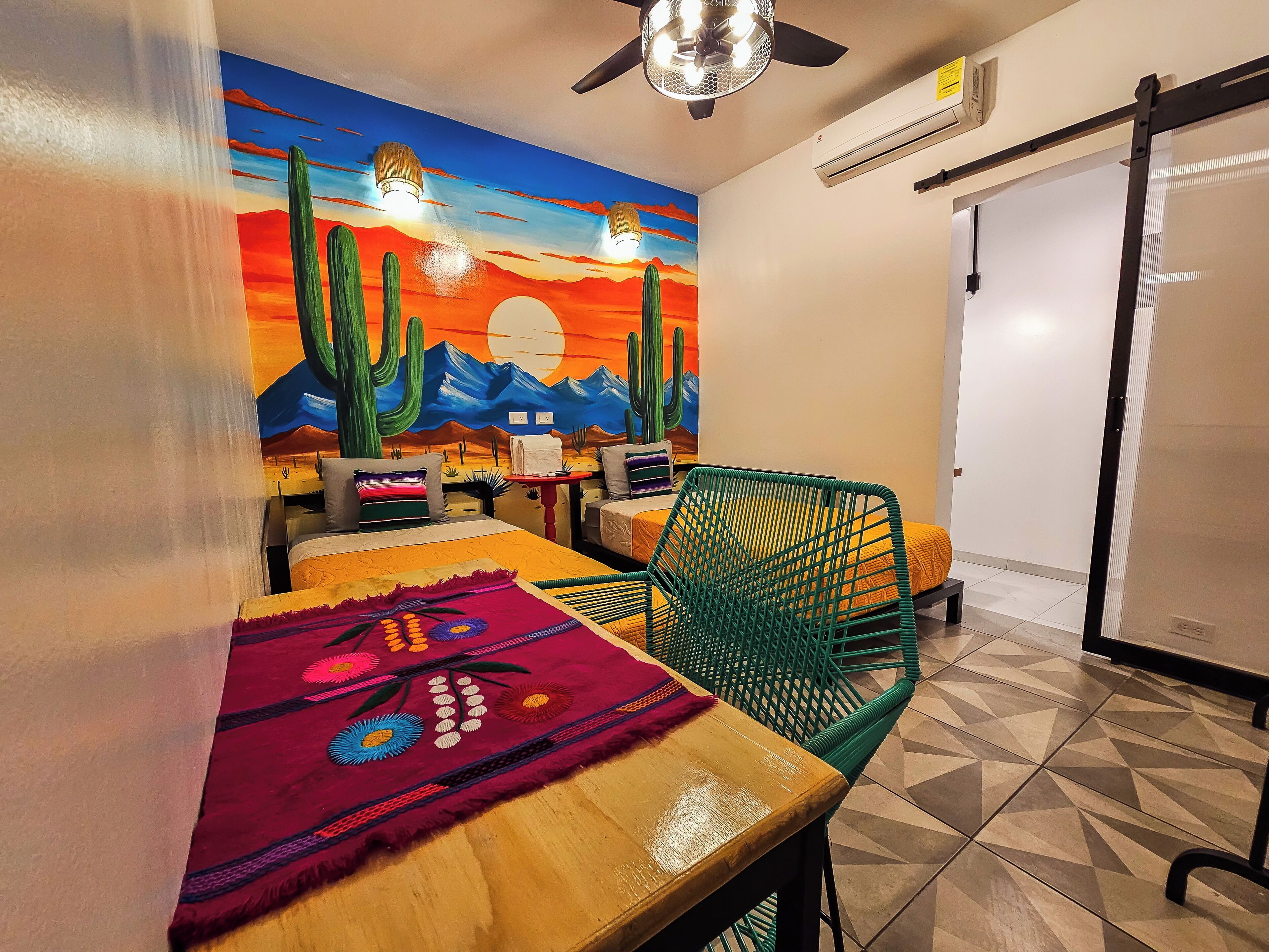 A room with a bed, a table with a colorful table runner, a green chair, and a mural of a desert scene.
