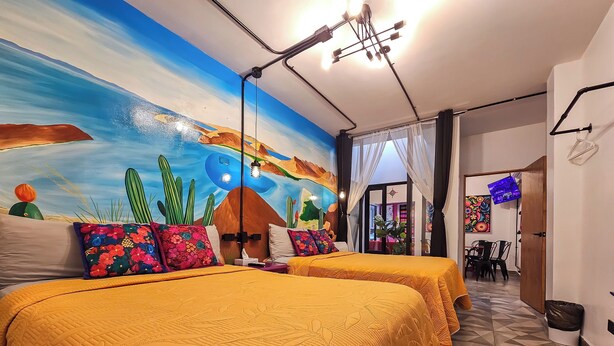 Ein Schlafzimmer mit zwei Betten, einem Wandgemälde und einem Essbereich, der durch eine offene Tür sichtbar ist.