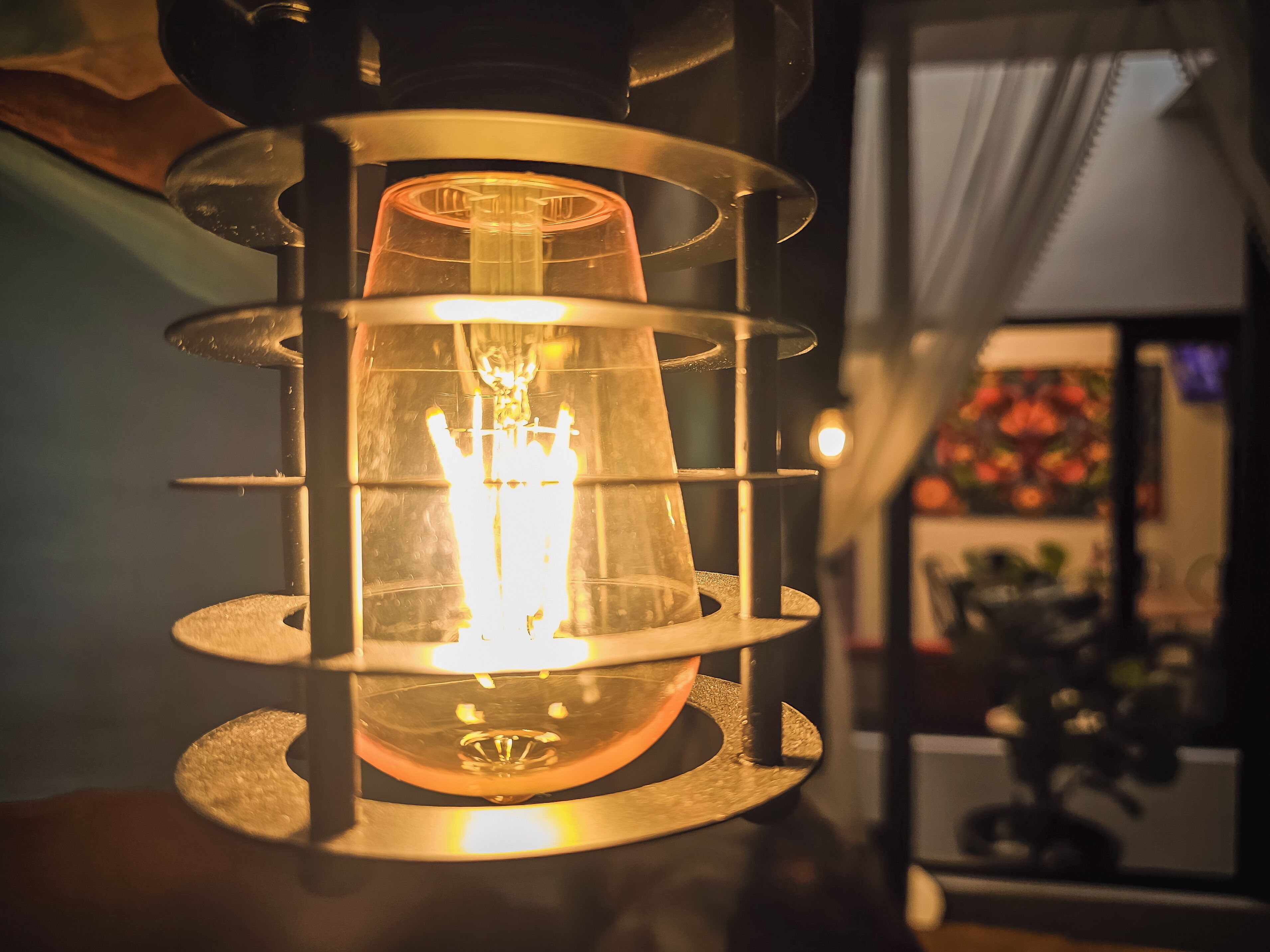 A multi-tiered, glass-encased lantern with a central light source, set against a blurred background with a window and a plant.