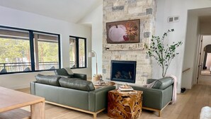 Una sala de estar moderna con chimenea de piedra, sofás verdes, una mesa de centro de madera y ventanas grandes.
