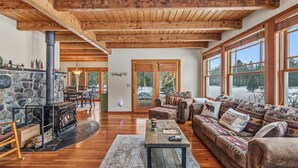 A spacious living room with a stone fireplace, wooden ceiling beams, and large windows offering a view of the outdoors.