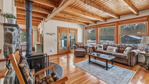 A spacious living room with a stone fireplace, wooden ceiling beams, and large windows offering a view of the outdoors.