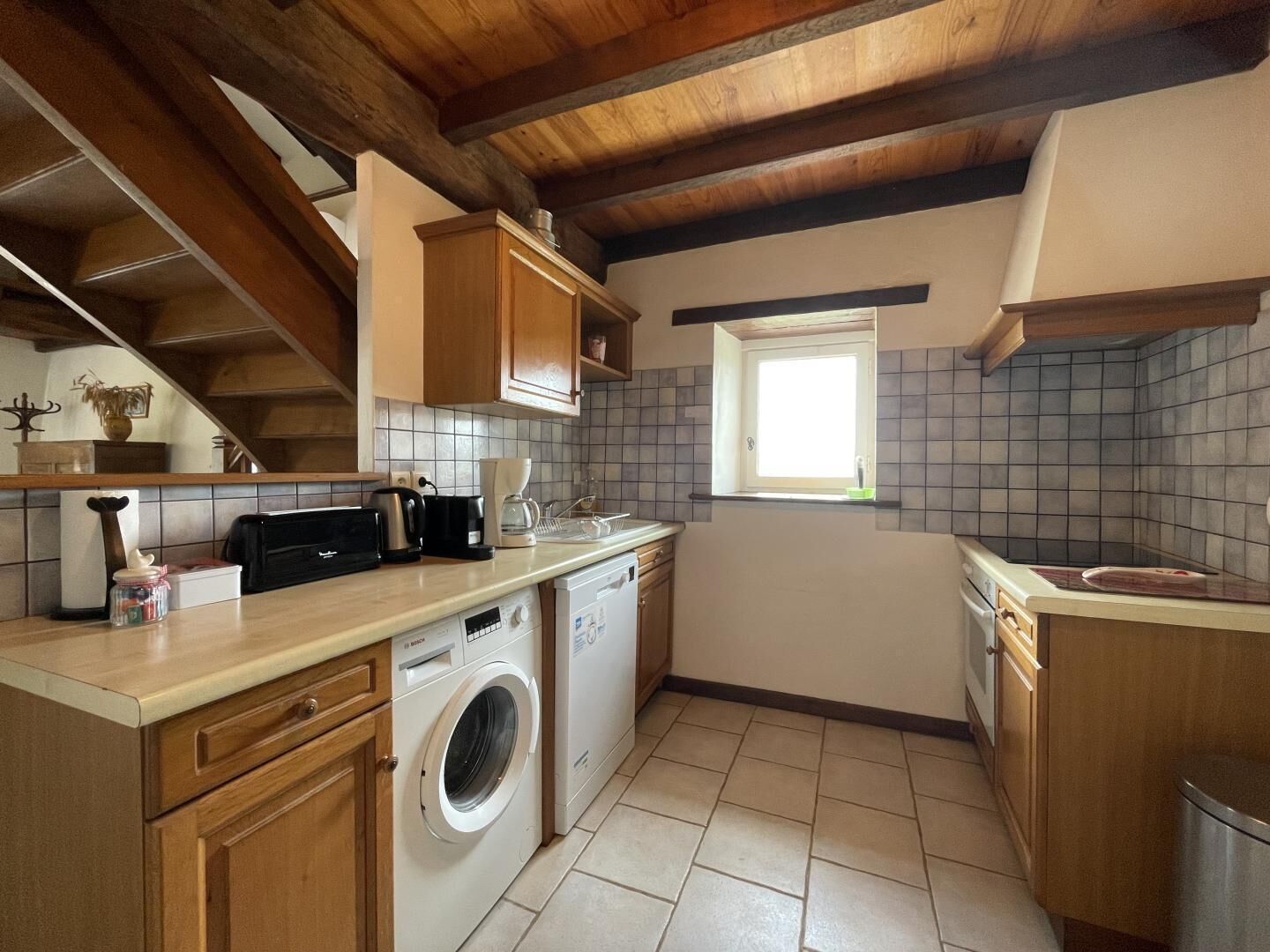 A compact kitchen with wooden cabinets, a washing machine, a coffee maker, and a window.