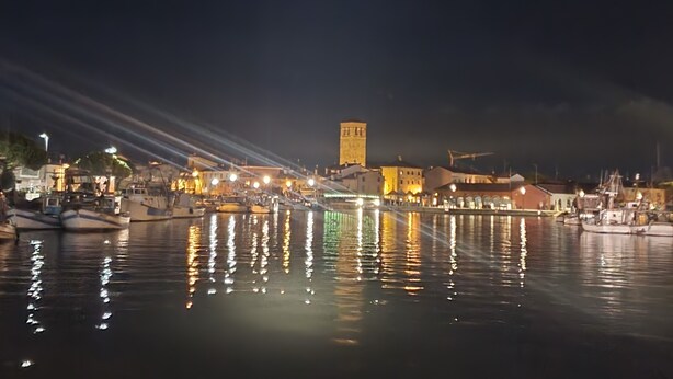 Una escena nocturna frente al agua con barcos amarrados, una torre destacada y edificios iluminados que se reflejan en el agua.