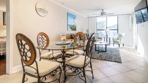 Un coin repas comprenant une table ronde en verre, quatre chaises à dossiers décorés, une horloge et un tableau représentant un phare.
