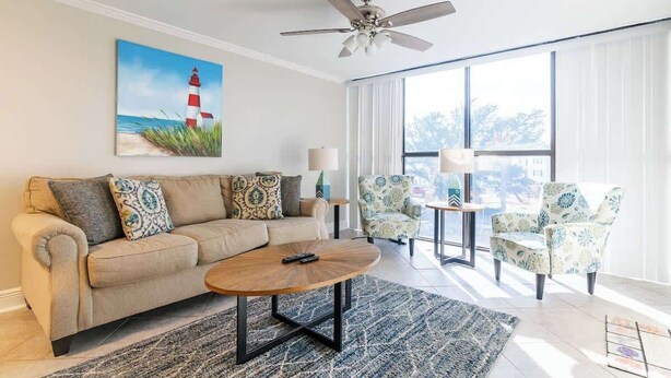 Un salon avec un canapé beige, des coussins à motifs, une table basse en bois et un tableau représentant un phare. On y trouve également deux fauteuils à motifs et une petite table avec une lampe.